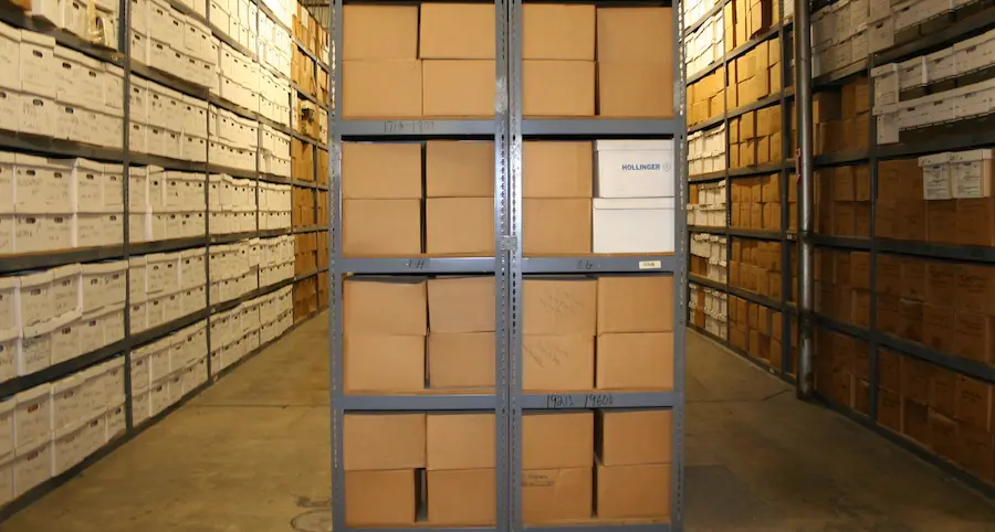 Here is a descriptive alt tag for this image: A long aisle in a large, secure archive storage facility, showing floor-to-ceiling metal shelving stacked with countless white and brown cardboard record boxes.