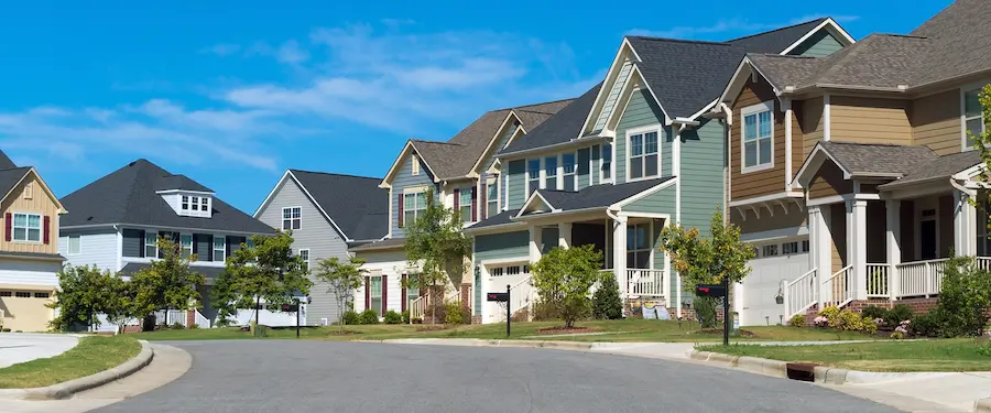 A row of modern, two-story suburban homes with varied colored siding and dark roofs, lining a curved residential street under a bright blue sky.