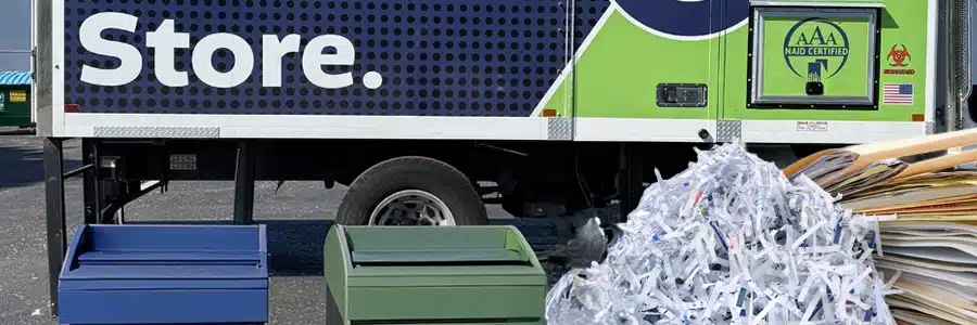 Side view of a mobile shredding truck displaying an AAA NAID Certified logo, parked behind two secure collection bins, a large mound of shredded paper strips, and a stack of file folders.