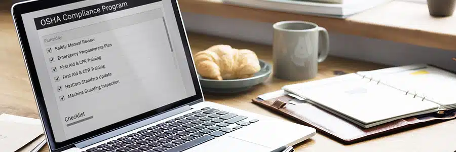 Close-up of a laptop screen displaying an "OSHA Compliance Program" checklist on a wooden desk, situated next to a coffee mug