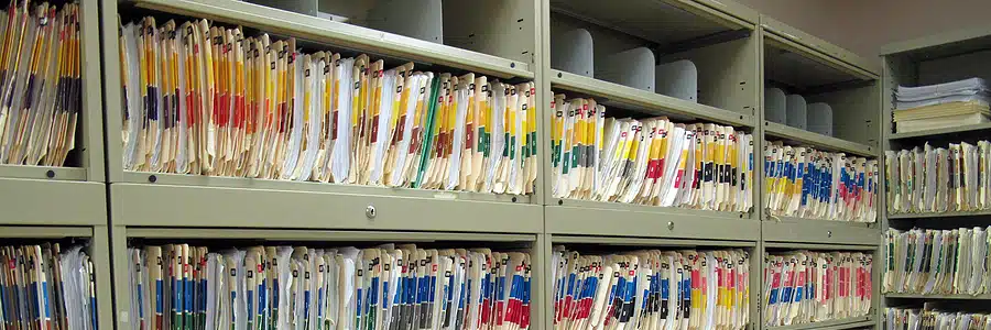 Shelves in a file room filled with color-coded hanging manila folders, representing a large volume of organized paper medical records or documents.