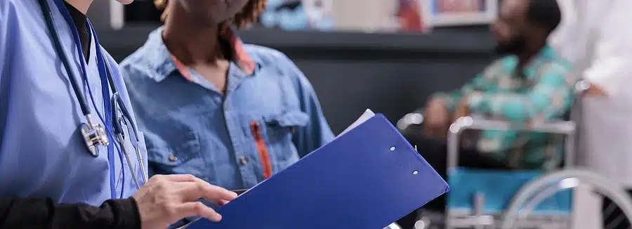 A medical professional is showing medical paperwork to a woman in a hospital while a man in a wheelchair waits in the background.